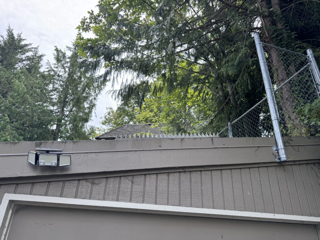 Außenansicht eines Gebäudedachs mit Ninja Deterrent-Schutzvorrichtungen, einschließlich Maschendrahtzaun, Kletterschutzspikes entlang der Kante und einer solarbetriebenen Bewegungsmelder-Leuchte zur Abschreckung von Eindringlingen.