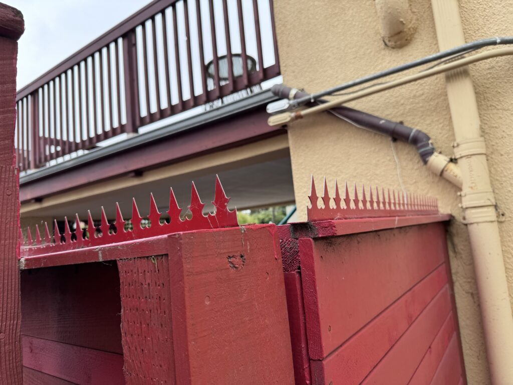 Clôture en bois rouge surmontée de pointes anti-escalade rouges, avec un garde-corps de balcon en arrière-plan.