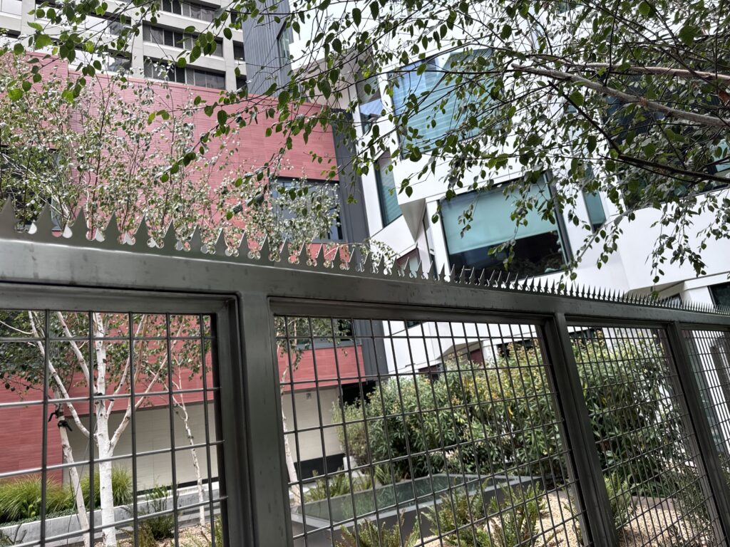 樹木とモダンな建物を背景にした、ステンレス製フェンススパイクのクローズアップ。