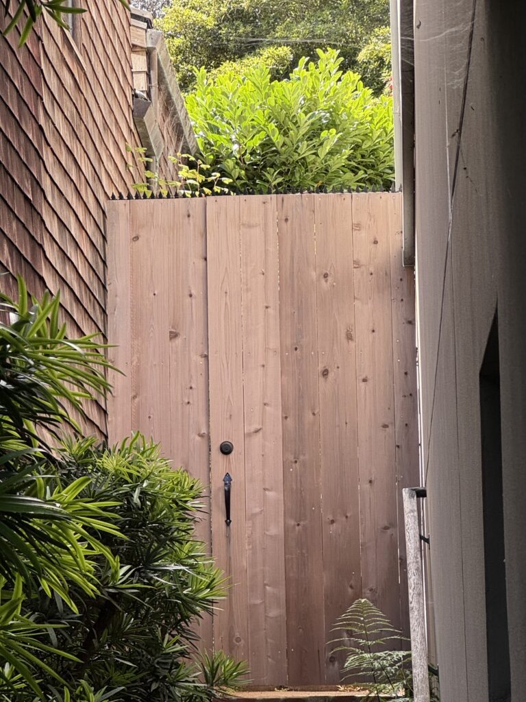 Portillon en bois au bout d’un passage bordé de verdure, avec des pics de sécurité anti-escalade installés sur le dessus.