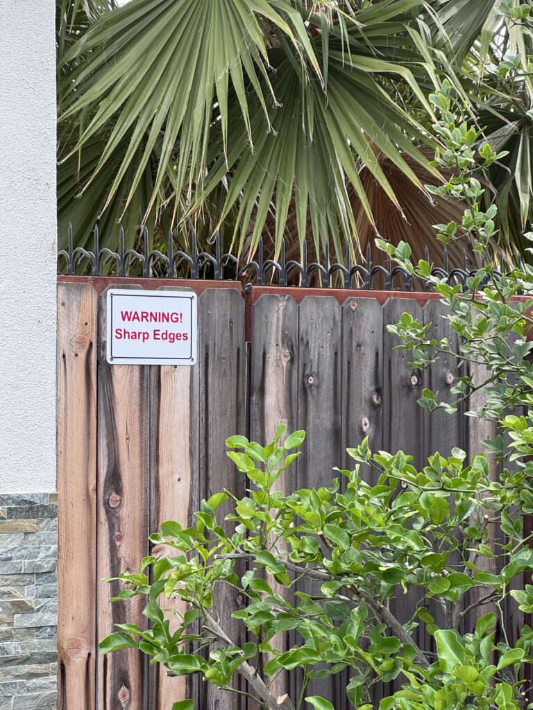 Close view of the wooden fence and “WARNING! Sharp Edges” sign, with black anti-climb spikes along the top and leafy branches in front.