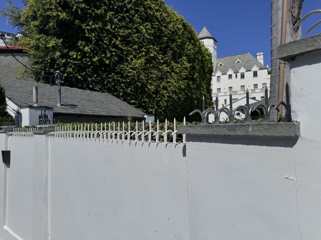White stucco perimeter wall topped with metal anti-climb spikes, with a large tree and a castle-like building in the background.