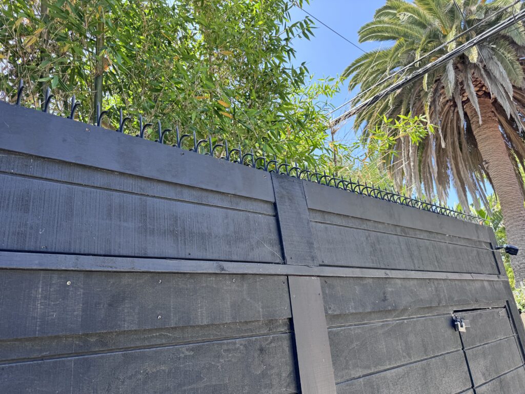 Angled view of the black wooden gate and latch detail, with curved security spikes and greenery overhead.
