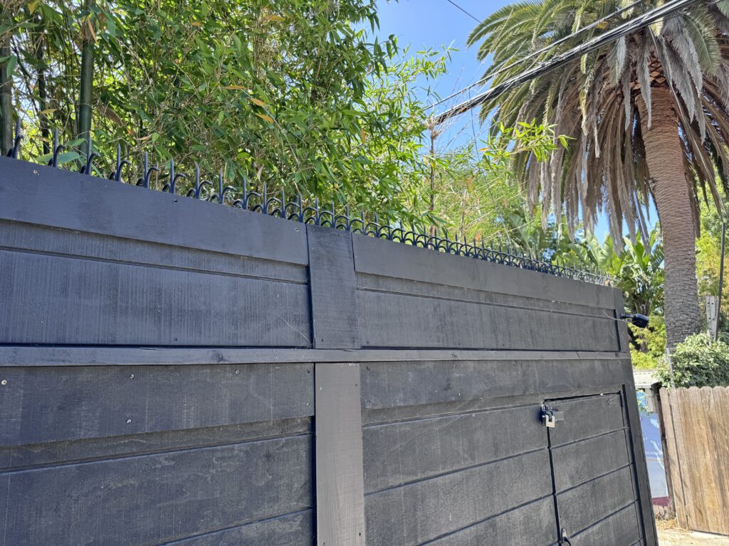 Corner view of a black wooden fence and gate, showing a continuous line of security spikes with trees behind.