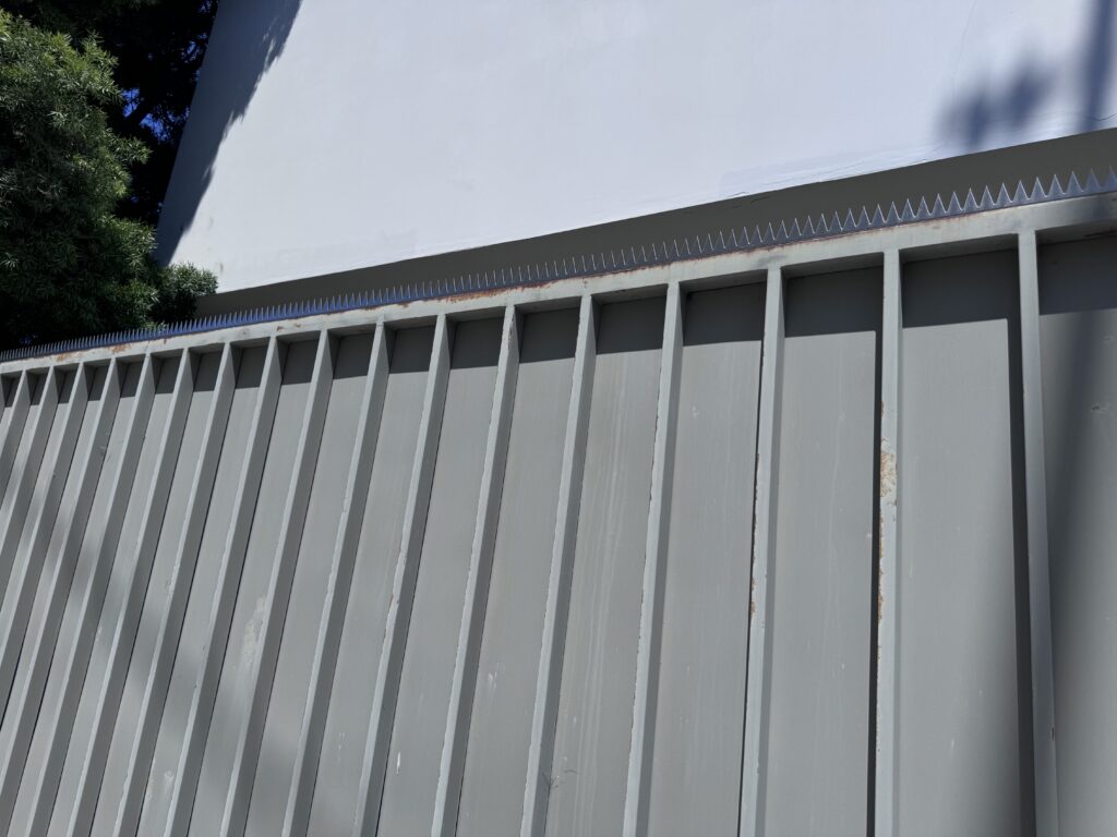 Long gray metal fence along a white building, with tree shadows stretching across the panels.