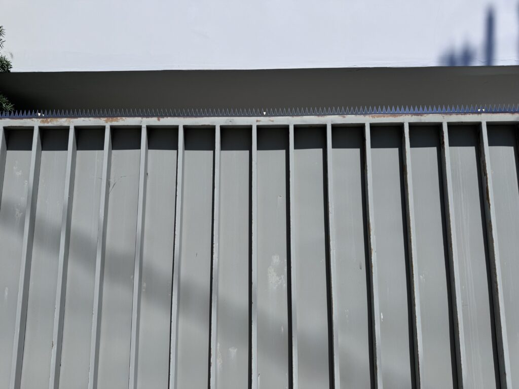Close view of the fence top showing a continuous strip of security spikes, with the white overhang casting shadows.