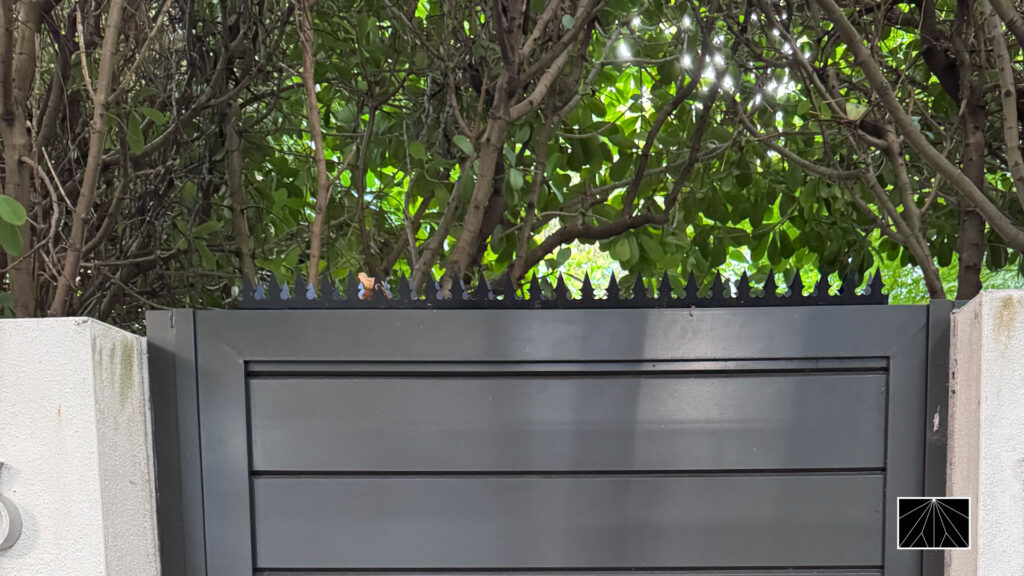 Black metal privacy gate with anti-climb finials along the top, set between concrete walls and shaded by dense greenery.