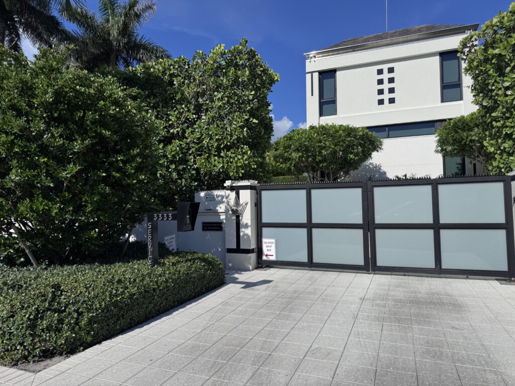 Casa moderna blanca detrás de un portón corredizo de vidrio esmerilado con marco negro, rodeada de árboles y setos bajo un cielo azul despejado.