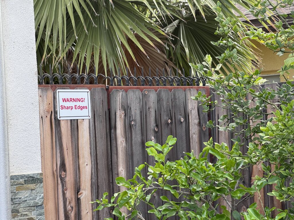 Valla de madera envejecida con púas antiescalada negras y un cartel “WARNING! Sharp Edges”, enmarcada por hojas de palmera y arbustos verdes.