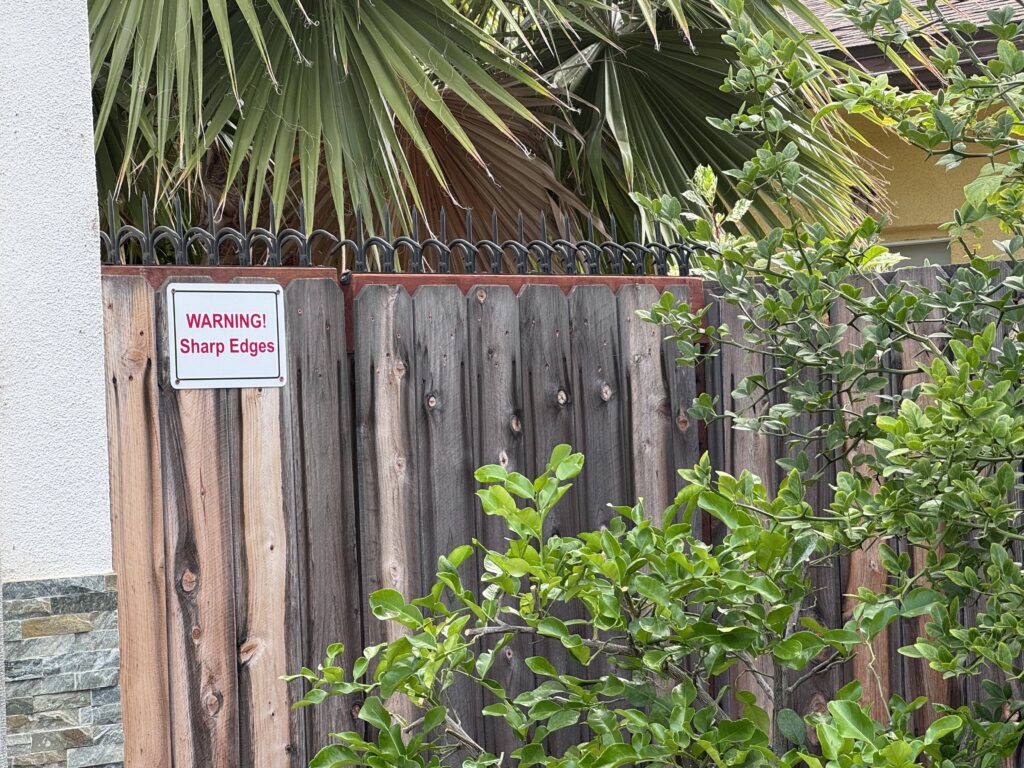 Gros plan de la clôture et du panneau « WARNING! Sharp Edges », avec pointes noires en partie haute et branches feuillues au premier plan.