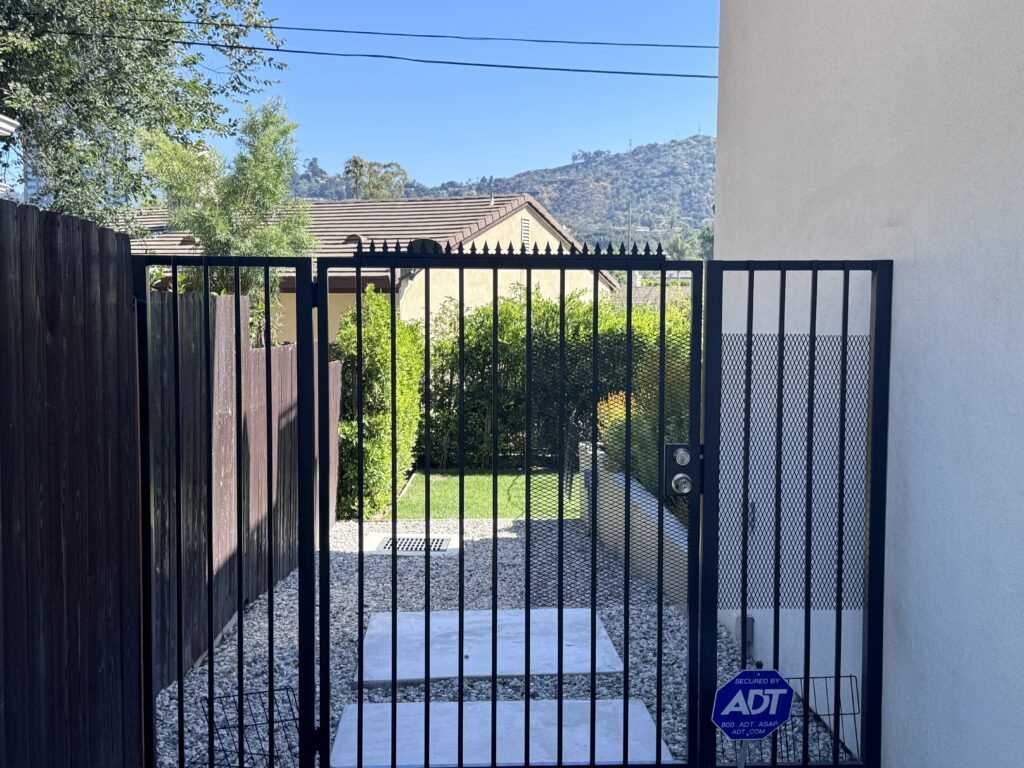 Portão lateral metálico preto com pontas anti-escalada e barras verticais, abrindo para um caminho de cascalho e um pequeno gramado, com colinas ao fundo.