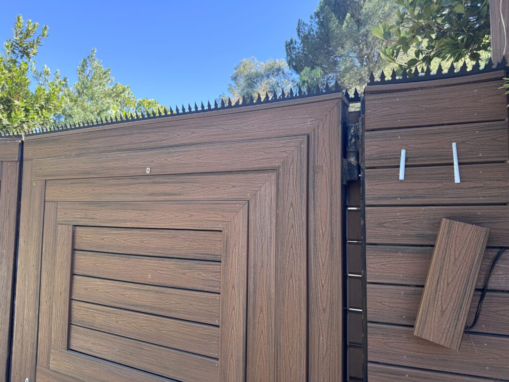 Portón de privacidad en acabado tipo madera con púas de seguridad anti-escalada negras instaladas en el borde superior bajo un cielo azul despejado