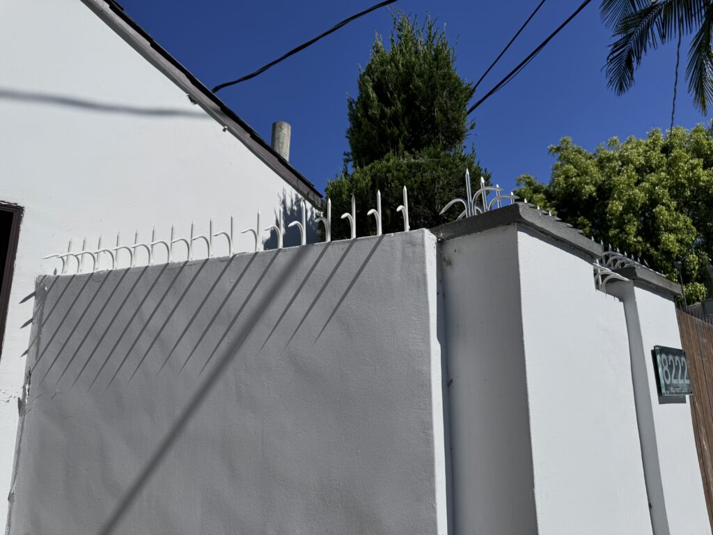 Vista en ángulo de una esquina de muro blanco con púas antiescalada proyectando sombras largas bajo un cielo azul profundo.