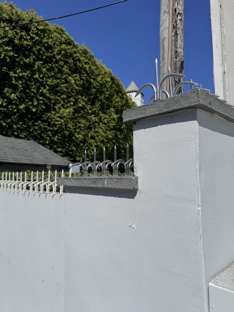 Close de um pilar de muro branco com pontas anti-escalada prateadas, com árvores e uma pequena torre ao longe.
