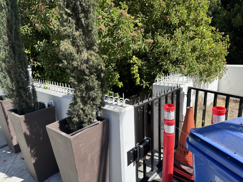 Área de entrada com um pequeno portão preto ao lado de um muro branco com pontas anti-escalada; vasos/planters e cones de segurança ao lado.