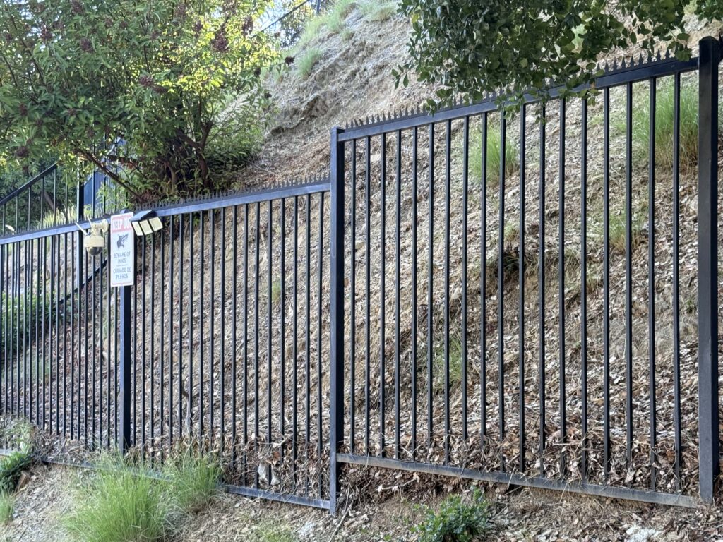 Cerca perimetral metálica negra con puntas anti-escalada en una ladera, con árboles y una señal de advertencia.