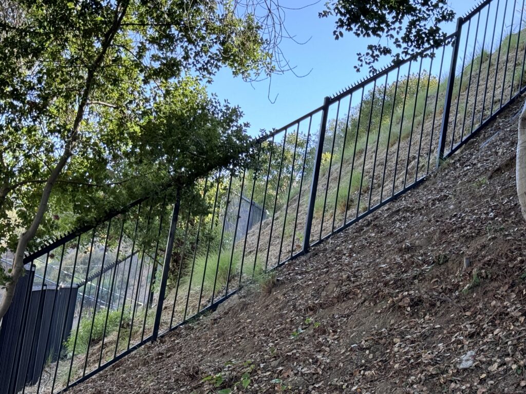 Cerca metálica negra con puntas anti-escalada a lo largo de una ladera pronunciada bajo árboles.