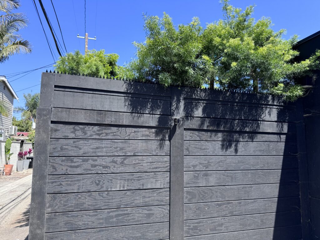 Portão de entrada em madeira preta com tábuas horizontais, com pontas antiescalada no topo sob céu azul limpo.