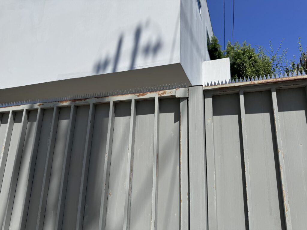 Eckdetail von grauem Tor und weißer Wand, mit einer Reihe Anti-Kletter-Spikes unter blauem Himmel und Stromleitungen.