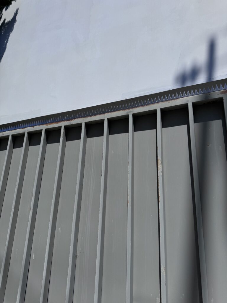 白い建物沿いに伸びる長いグレーのフェンス。パネル上に木陰が伸びている。