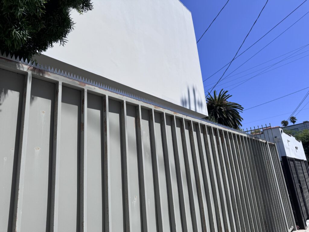 Vista en ángulo de una valla metálica gris con nervaduras verticales bajo un muro blanco, cielo azul y cables eléctricos arriba.