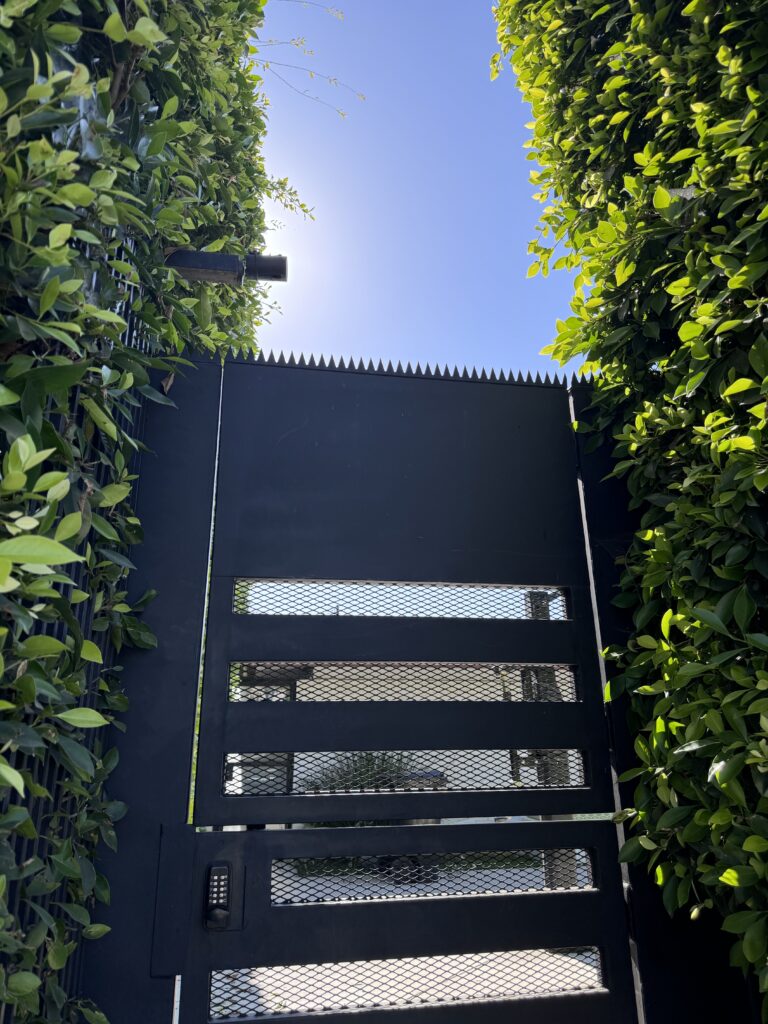 Schwarzes Metalltor zwischen hohen Hecken, mit Anti-Climb-Spikes obenauf, mit Gitterpaneelen und Codeschloss.