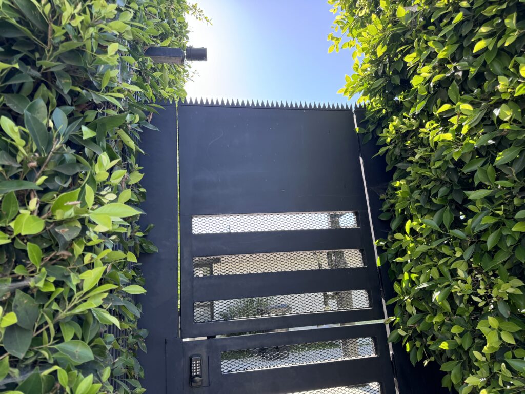 Portão de segurança metálico preto com painéis de malha e pontas anti-escalada, emoldurado por sebes densas.