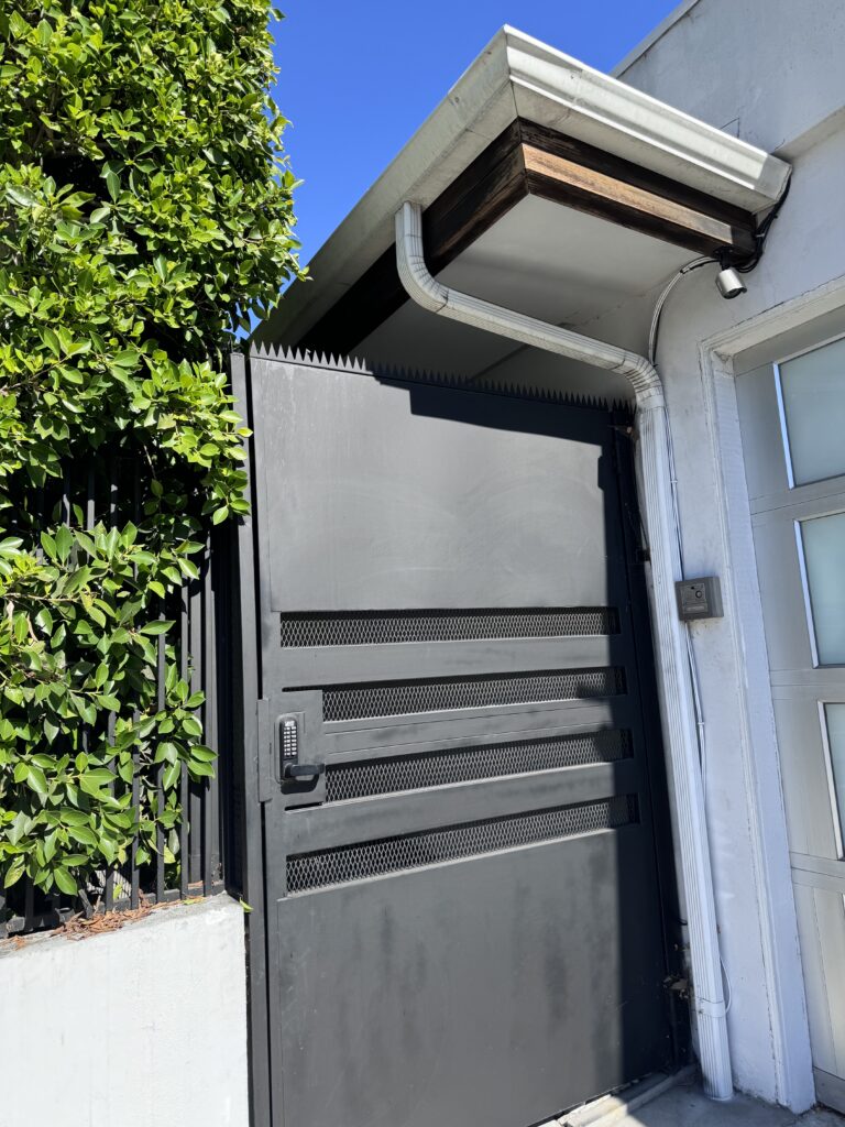 Entrada lateral: portão metálico preto com fechadura com teclado e pontas anti-escalada, ao lado de um edifício branco.