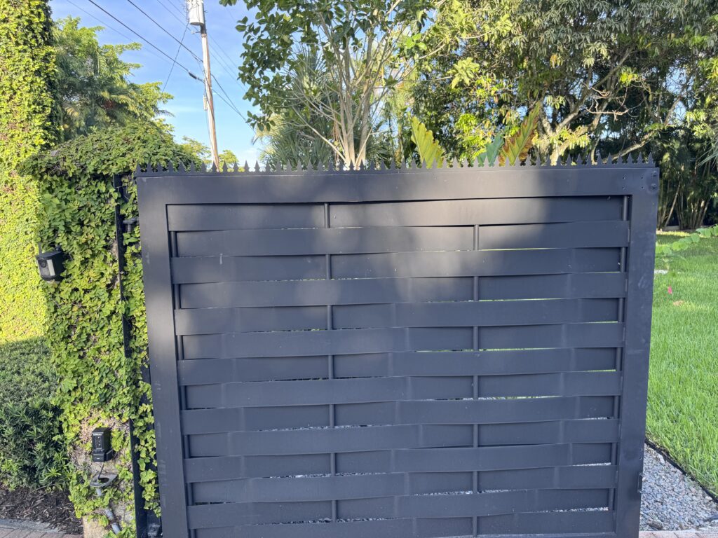 Vista amplia a través de portones negros abiertos hacia una gran casa blanca, palmeras altas y una entrada curva de adoquines