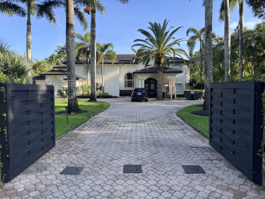 Vista amplia a través de portones negros abiertos hacia una gran casa blanca, palmeras altas y una entrada curva de adoquines