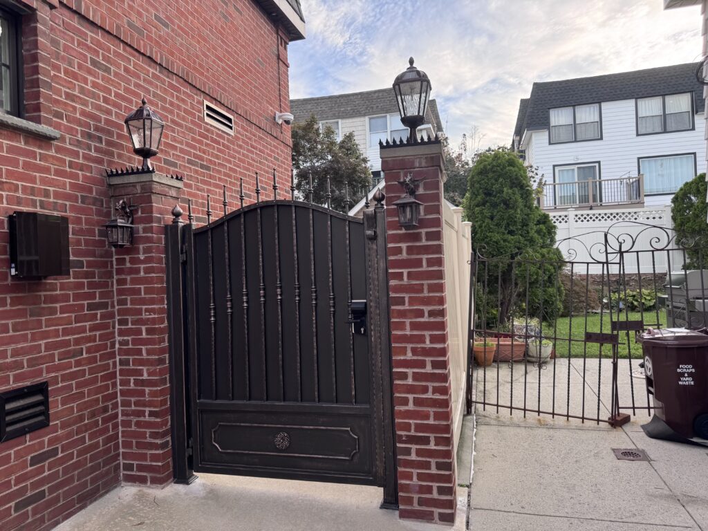 주택 드라이브웨이에서 레드 브릭 기둥 사이에 놓인 검은 금속 보행자 게이트, 상단의 안티클라임 스파이크와 랜턴 조명