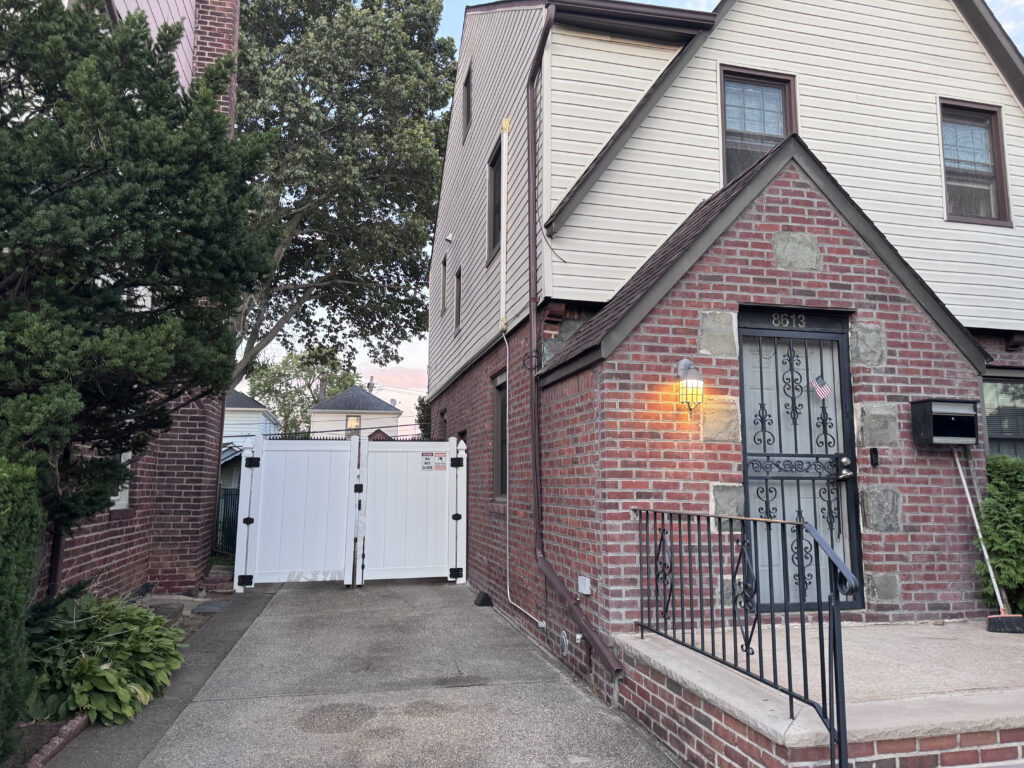 Extérieur de maison en brique avec porte d’entrée en fer forgé et marches, menant à une allée avec portail latéral blanc