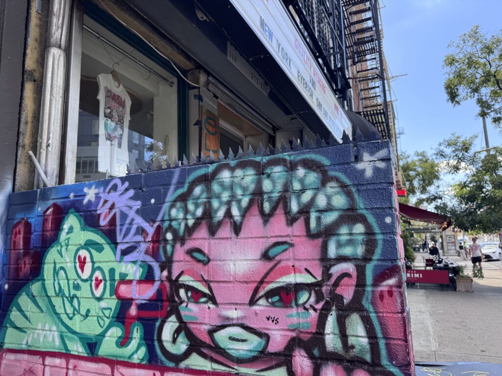 Mur de brique recouvert de graffiti, surmonté de pointes anti-escalade devant une devanture new-yorkaise, avec trottoir et escalier de secours visibles